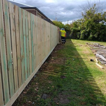 A worker in high-visibility clothing installing a new timber paling fence along a residential backyard, with old fence materials stacked on the grass.